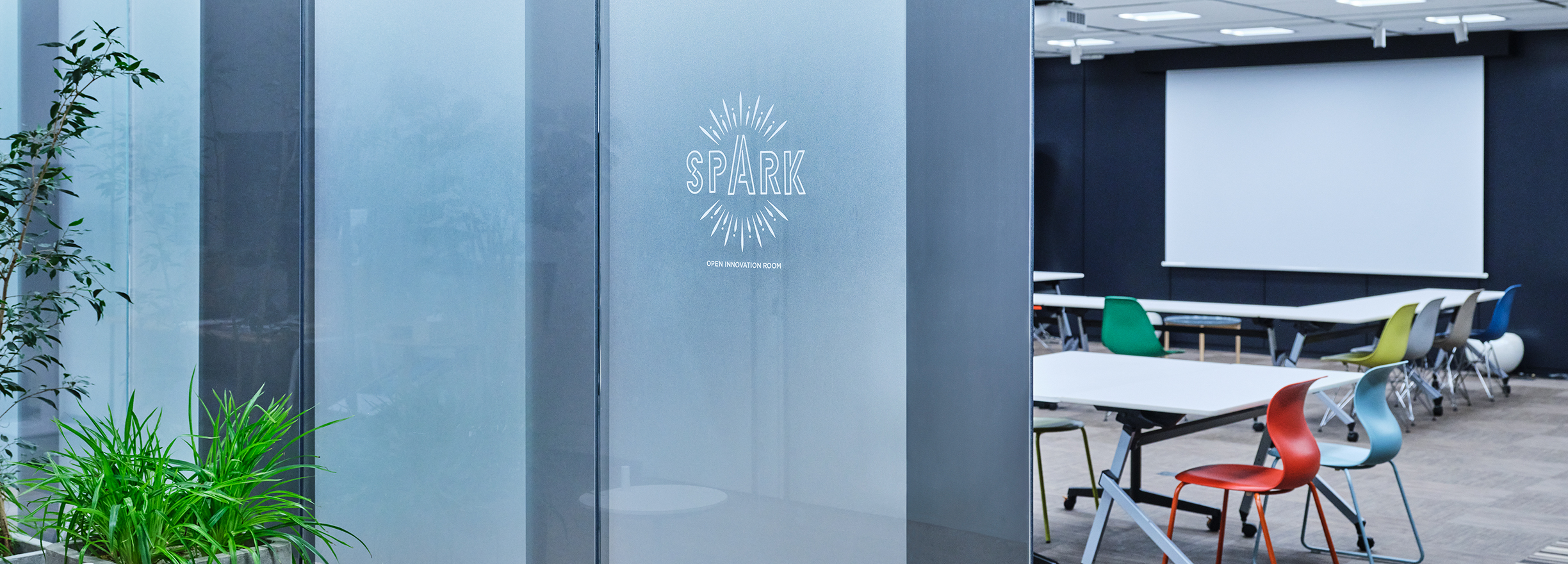 モダンなオフィススペースの入り口。ガラスのドアに「SPARK」のロゴが描かれている。内部には会議室やワークスペースが見える。カラフルな椅子とプロジェクタースクリーンが設置されている。入り口付近には観葉植物が置かれている。